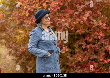 A woman stands confidently in a denim coat and a stylish hat, smiling softly as she poses beside vibrant red and orange foliage in a park during autum Stock Photo