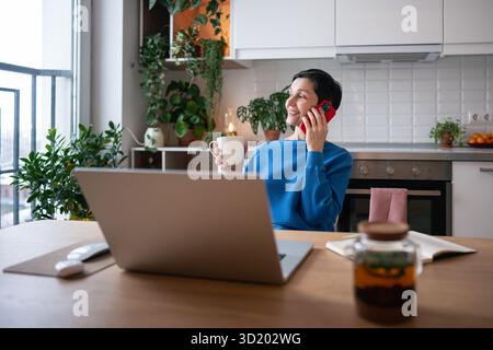 Smiling mature woman enjoy phone call and herbal tea. Relaxed comfortable home job with slow living Stock Photo