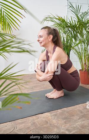A woman squats on a colorful yoga mat in front of potted plants Stock Photo