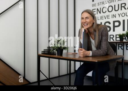 Professional woman smiling while seated at a desk in a modern office with motivational quotes on the wall Stock Photo