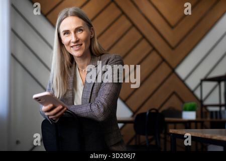 Professional woman in modern office environment using smartphone while sitting on chair with stylish wooden backdrop Stock Photo