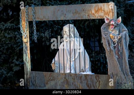 Spooky Halloween display featuring a skeleton ghost figure in a wooden jail cage next to a creepy vampire bat creature with chains and eerie lighting Stock Photo