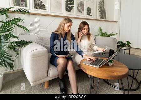 Two women collaborating on a laptop while sitting on a sofa in a modern, plant-filled office space Stock Photo