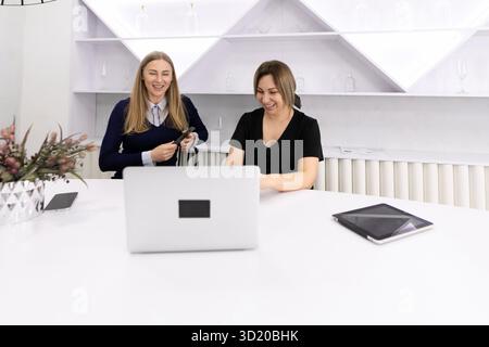 Two women collaborating at a modern workspace, engaging in a discussion while using a laptop and tablets, showcasing teamwork an Stock Photo