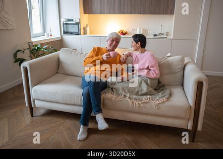 Happy laughing mature couple engaged in lively conversation, drinking tea on cozy sofa together Stock Photo