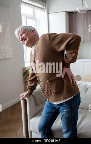 Suffering elderly man hunched with back pain struggles to rise from couch due to spinal problems Stock Photo