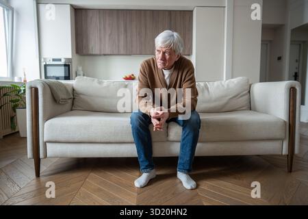 Lonely abandoned elderly man sits lost in thought on sofa with crossed arms, depressed memory loss Stock Photo
