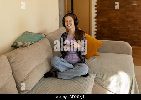 Young woman enjoys gaming while wearing headphones in cozy living room setting during afternoon Stock Photo