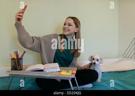 Young woman takes a selfie while studying at her cozy home workspace with a cute dog nearby Stock Photo
