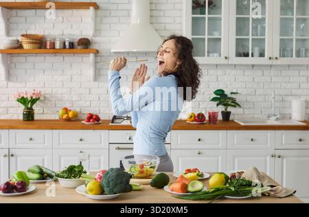 Joyful young woman singing in kitchen and using spatula as microphone while preparing fresh salad. Stock Photo