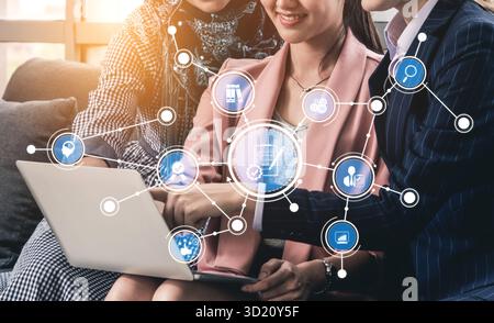 Three business professionals collaborate on a laptop, discussing innovative ideas with digital connectivity icons, symbolizing teamwork and modern tec Stock Photo