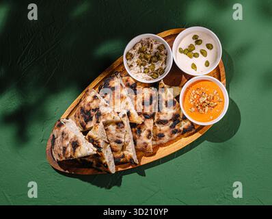 Delicious freshly baked flatbread served with assorted dips on a wooden platter against a green background Stock Photo