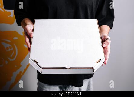 Individual holding a blank white pizza box in a casual setting with a colorful wall background Stock Photo