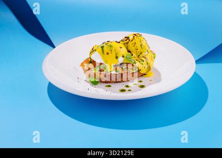 Delicious eggs benedict served on toast with vibrant yellow sauce and green garnish against a bright blue backdrop Stock Photo