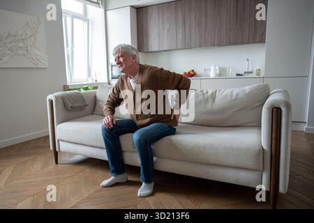 Sad depressed senior man suffering from osteochondrosis barely stand up from sofa, acute back pain Stock Photo