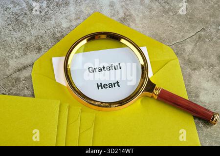 A paper with the text Grateful Heart can be seen through a magnifying glass on top of a stack of yellow envelopes, all lying on a grey stone surface. Stock Photo