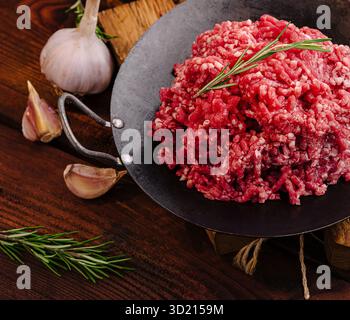 Fresh ground beef rests in a black skillet surrounded by garlic cloves and rosemary sprigs on a rustic wooden table, highlighting ingredients for flav Stock Photo