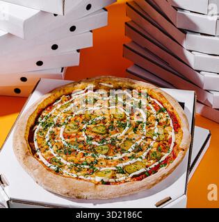 Delicious pizza topped with vegetables and herbs placed in front of stacks of pizza boxes in a vibrant setting Stock Photo