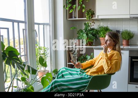 Pensive woman staring out window holding phone in plant filled kitchen. Lost in thoughts, bad news Stock Photo