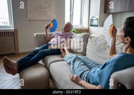 Stressed elderly couple suffering from heat, fanning on sofa unable to function. Seniors struggle Stock Photo