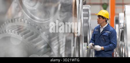 Industrial worker in blue uniform and yellow safety helmet inspecting machinery at factory, engineer technician monitoring and gears background wide f Stock Photo