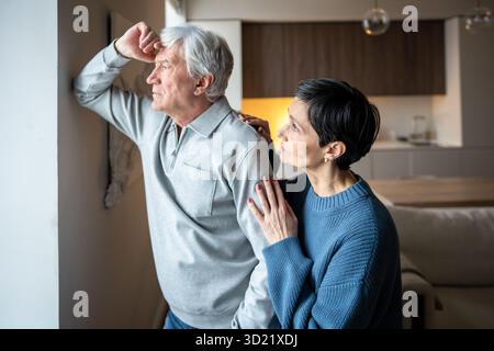 Thoughtful sad senior man look out window while wife reach out, consoling. Family support, empathy Stock Photo