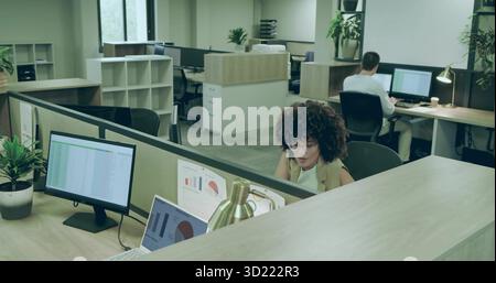 Curly-haired worker in sleeveless top reviewing charts on monitor at desk with lamp, copy space Stock Photo