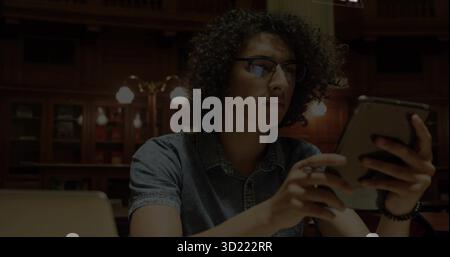 Studying male in denim shirt using handheld tablet at library study table, with laptop, desk lamp Stock Photo