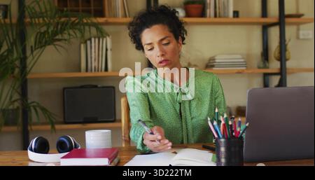 Writing mature woman wearing green blouse at home office desk, using pen with laptop and headphones Stock Photo