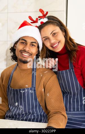 Diverse couple standing in home kitchen wearing striped aprons and festive headwear with flour dust Stock Photo