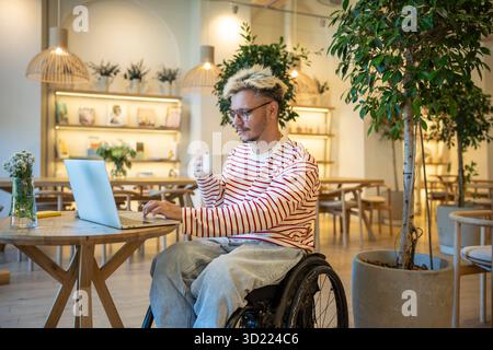 Focused guy freelancer with disabilities in wheelchair working online using laptop sits in cozy cafe Stock Photo