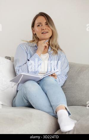 Woman with thoughtful expression fills diary sitting on sofa Stock Photo
