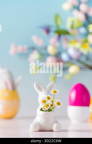 Easter card. A white bunny with a bouquet of daisies on a blue background and colored eggs. Stock Photo