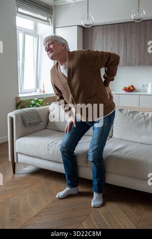 Struggling old man with severe back pain struggles to rise from couch at home due to spinal problems Stock Photo