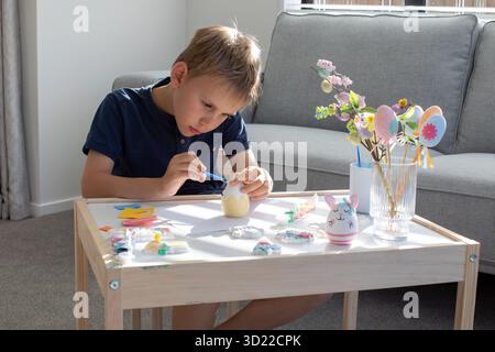 A boy is sitting at a table and decorating an Easter egg. The concept of Easter children's crafts. Stock Photo