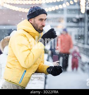 Middle aged man drinking hot coffee or tea from cup on ice rink outdoor in snowy winter day Stock Photo