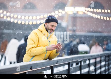 Middle aged man standing near fence on ice rink and using smartphone, outdoors at snowy day Stock Photo