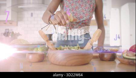 Woman in sleeveless top squeezing lemon half over greens at kitchen countertop, with salad bowl Stock Photo