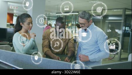 Pointing at keyboard three colleagues discussing project at cubicle desk in office, with data icons Stock Photo