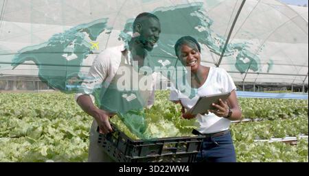 Two farm workers monitoring lettuce trays in greenhouse, holding crate and tablet with map overlay Stock Photo