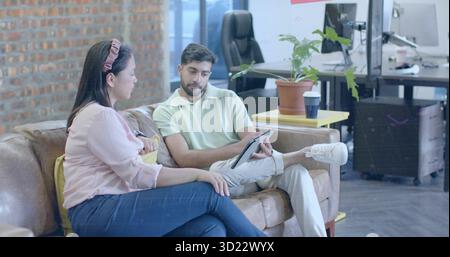 Reviewing tablet content by two coworkers seated on sofa in office lounge, with potted plant Stock Photo