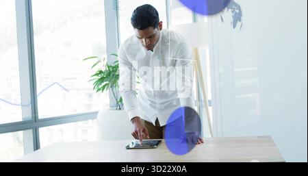 Leaning man in white shirt, brown trousers using tablet at desk in office with data overlays Stock Photo