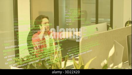 Talking Asian man holding smartphone, pointing at screen across cubicle partition in office Stock Photo