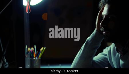 Leaning forehead on palm man contemplating work at home study, with desk lamp and colored pencils Stock Photo