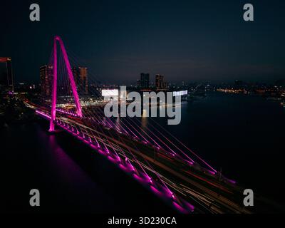 Aerial view of a brightly lit pink bridge spanning a large river during nighttime in an urban city. Ho Chi Minh City, Vietnam Stock Photo