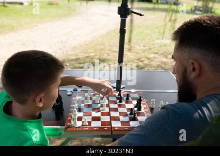 Father and son engage in a friendly game of chess on a portable board set up outside. The relaxed setting suggests leisure time spent together. Stock Photo