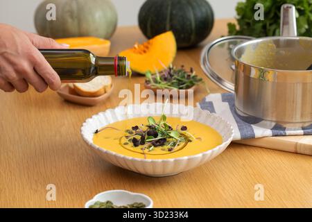 Female hand pouring olive oil into pumpkin soup with microgreens garnish on wooden table Stock Photo
