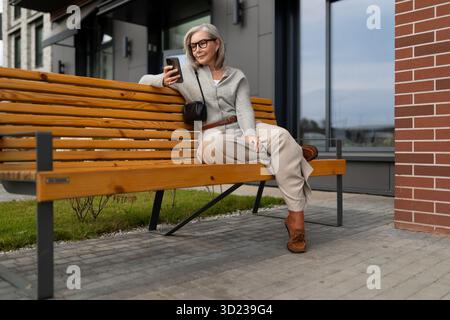 Elderly woman relaxing on a bench using her smartphone in an urban outdoor setting during daytime Stock Photo