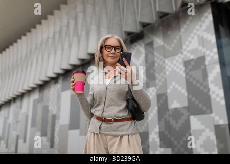 Senior woman enjoying coffee while using smartphone outside a modern building in an urban setting Stock Photo
