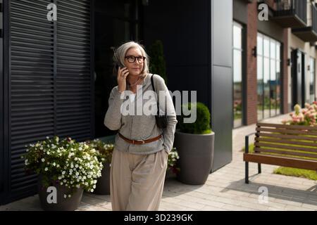 Mature woman walking along a modern street while talking on her phone in a pleasant neighborhood setting Stock Photo